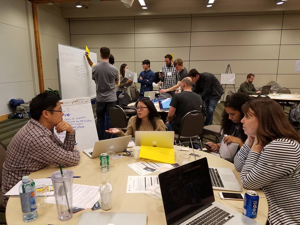 Four people sitting around the table in the foreground discussing a project while others work at a whiteboard in the background